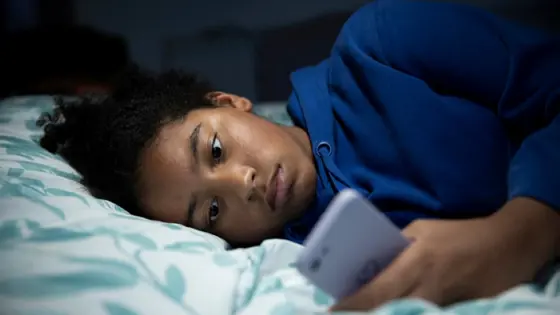 A young person lying in bed on their phone. The lights are off, they're meant to be asleep.